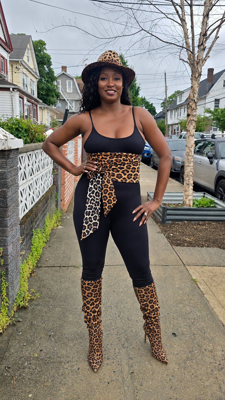 Leopard print belt styled over a black jumpsuit with matching hat and knee-high boots